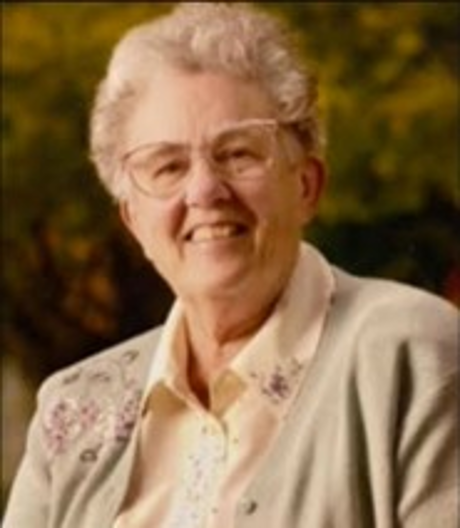 A portrait of Mary Buzzell, with curly grey hair, glasses, smiling at the camera